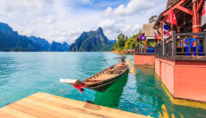 Een traditionele langstaartboot aangemeerd aan een houten steiger, met een zwerm vissen zichtbaar in het heldere meerwater. Drijvende accommodaties zijn in de buurt te zien en majestueuze bergen rijzen op de achtergrond op onder een bewolkte hemel in Khao Sok, Thailand.